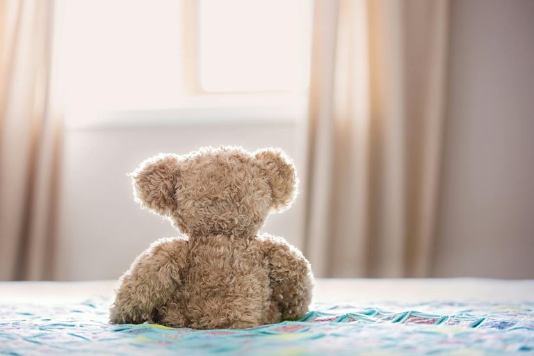 A plush teddy bear sits on a colorful bed in a cozy bedroom, facing a window.